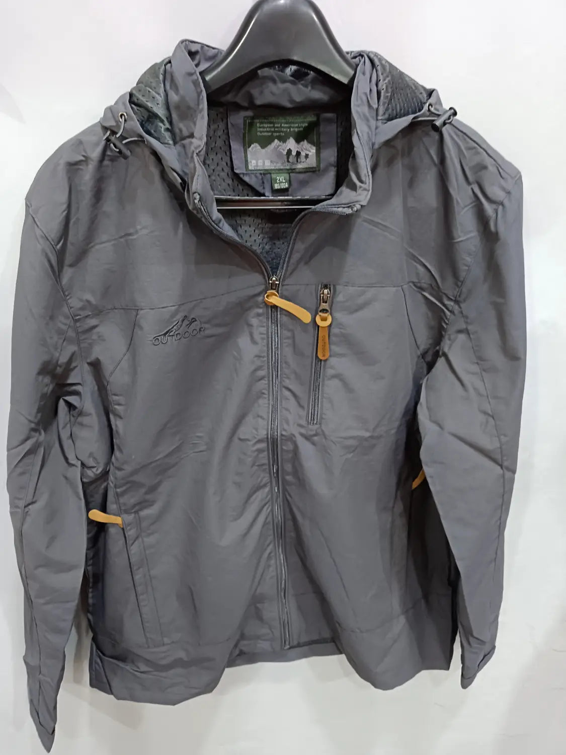 Chaqueta de senderismo con capucha impermeable en color gris, con tiradores de cremallera color camel y un logotipo de montaña sutil en el pecho, diseñada para actividades al aire libre.