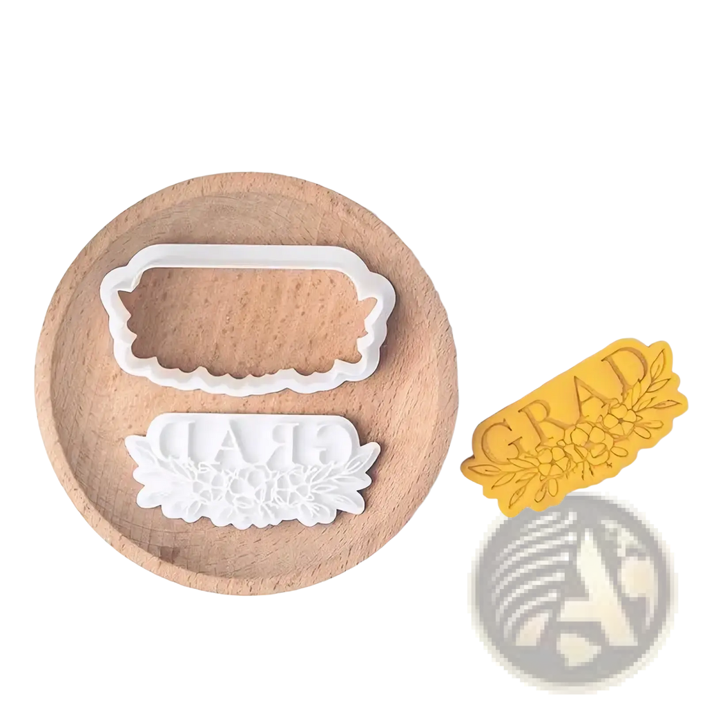 White plastic grad cookie cutter with floral design and yellow pressed dough sample from 2026 graduation cookie cutter set