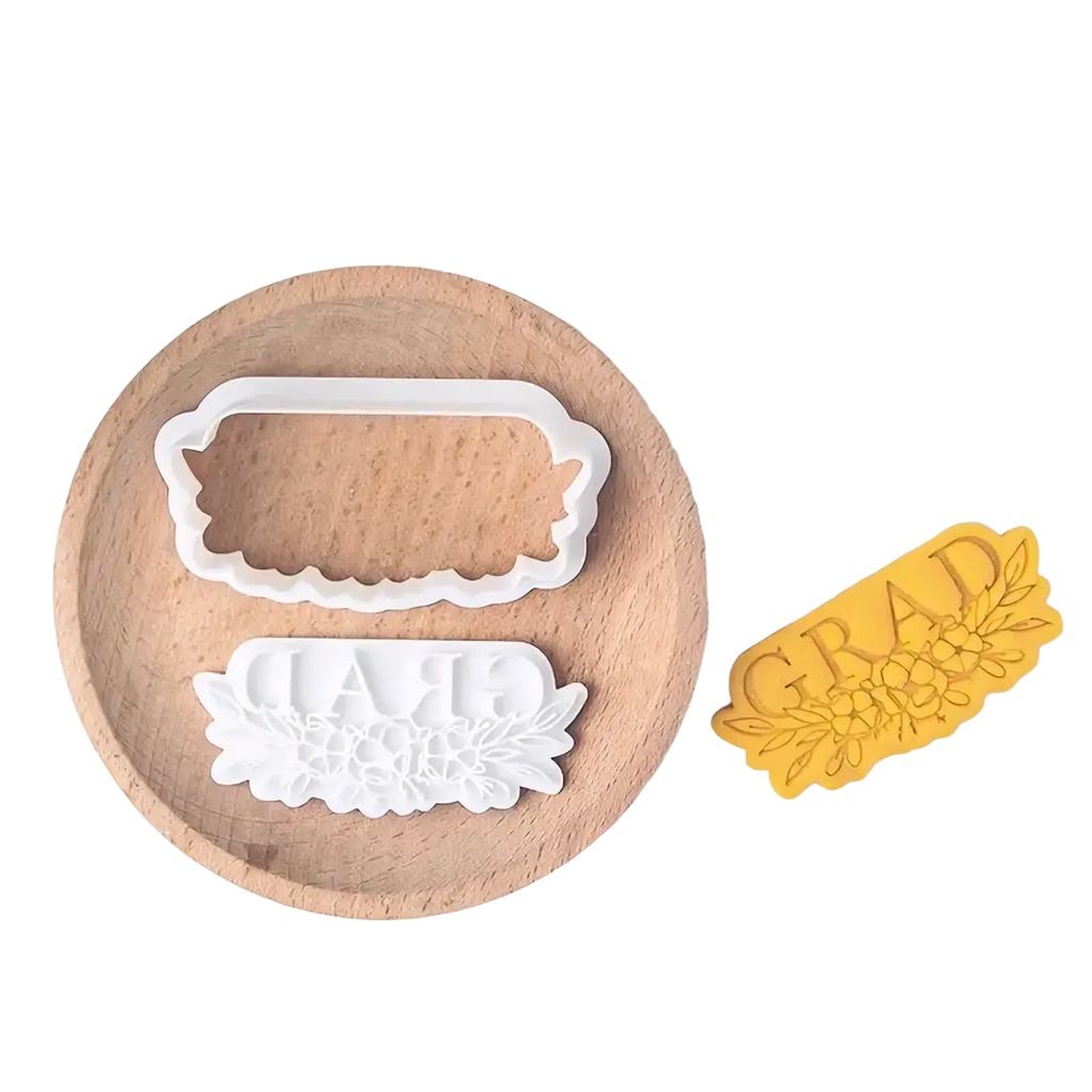 White plastic grad cookie cutter with floral design and yellow pressed dough sample from 2026 graduation cookie cutter set