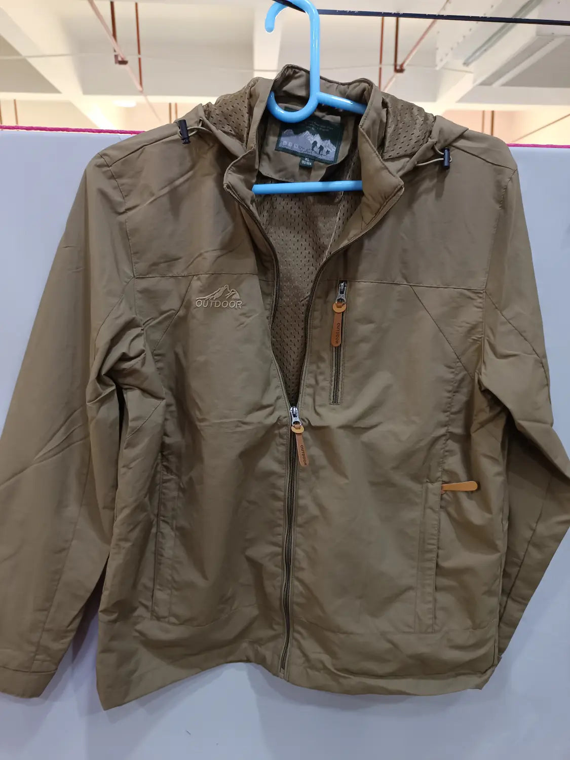 Chaqueta de senderismo con capucha impermeable color camel, con cierre frontal mediante cremallera y bolsillos laterales, diseñada para actividades al aire libre.