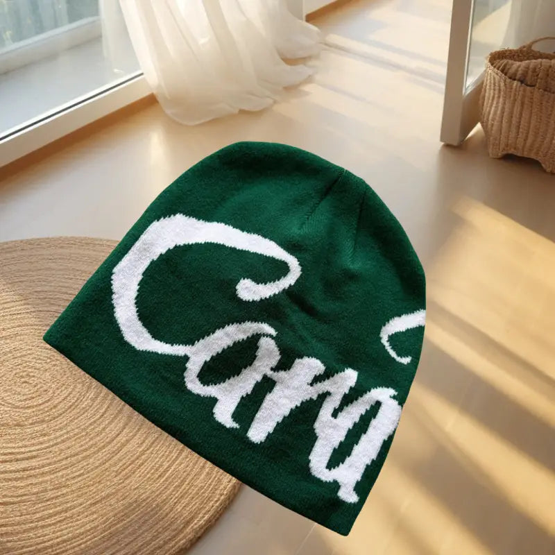 A dark green knitted beanie hat featuring a bold white cursive car logo embroidered across the front.