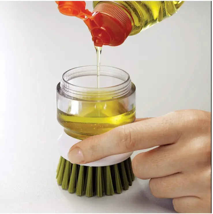 Soap dispensing dish brush with non slip storage base featuring green bristles, a clear plastic jar base, and a white handle being filled with yellow liquid soap from an orange dispenser.