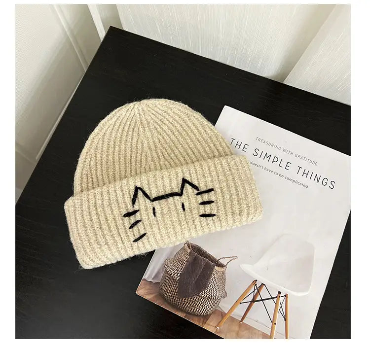 Cream-colored knitted beanie hat featuring a black embroidered cat face design on the folded brim.