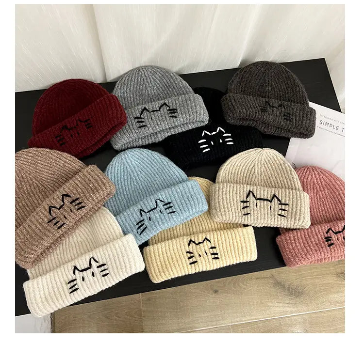 A collection of cozy knitted beanie hats in soft pastel and earthy tones featuring a simple embroidered cat face on the folded brim.