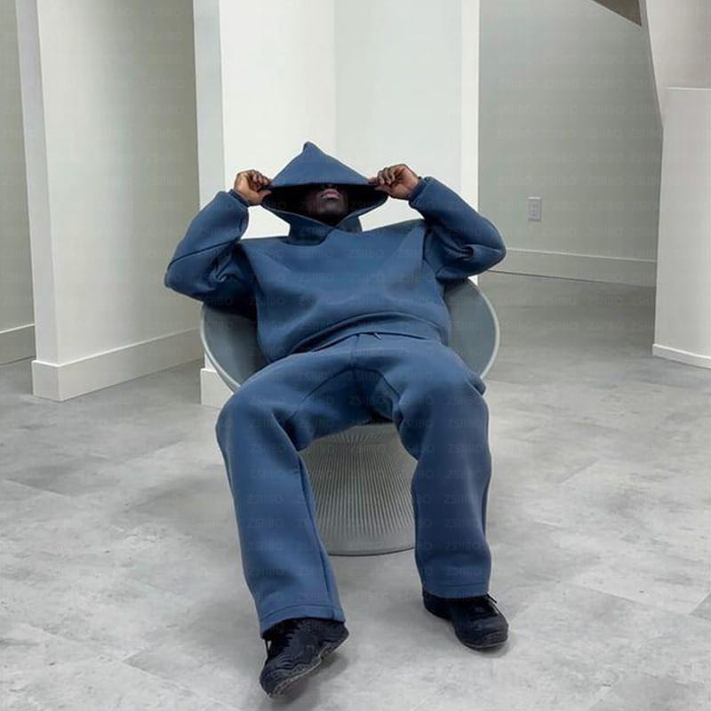 Une personne portant un ensemble oversize unisexe streetwear, sweat à capuche et pantalon de survêtement bleu marine, assise sur une chaise grise moderne.