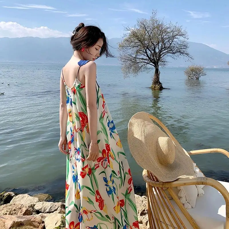 A woman wears a floral mesh maxi dress with a retro slip silhouette featuring long sleeves thin straps and vibrant red blue yellow and green flowers on an off-white background.