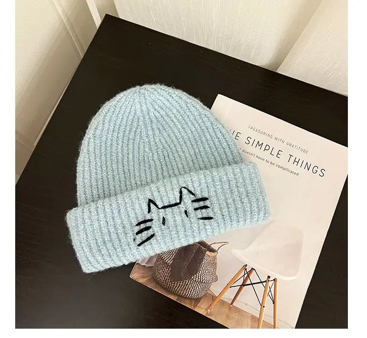 A soft light blue knitted beanie hat with a ribbed texture and a black embroidered cat face on the folded brim.