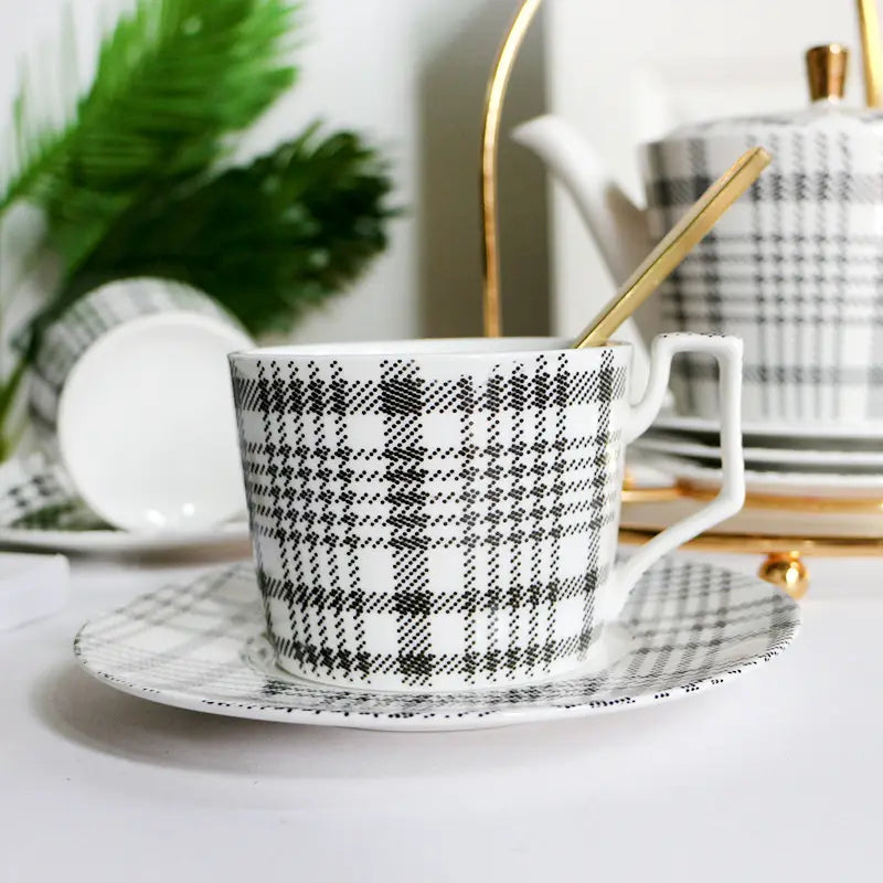 A white ceramic teacup with a black plaid pattern and gold-toned accents on a matching saucer from a ceramic tea set with plaid teapot and gold trim tray.