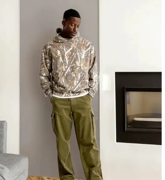 A man wearing an oversized leafy camouflage lantern sleeve hoodie in beige brown and white tones paired with olive green cargo pants.