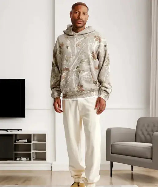 a man wearing an oversized leafy camouflage lantern sleeve hoodie in beige with an intricate floral pattern in muted earth tones paired with cream-colored joggers.
