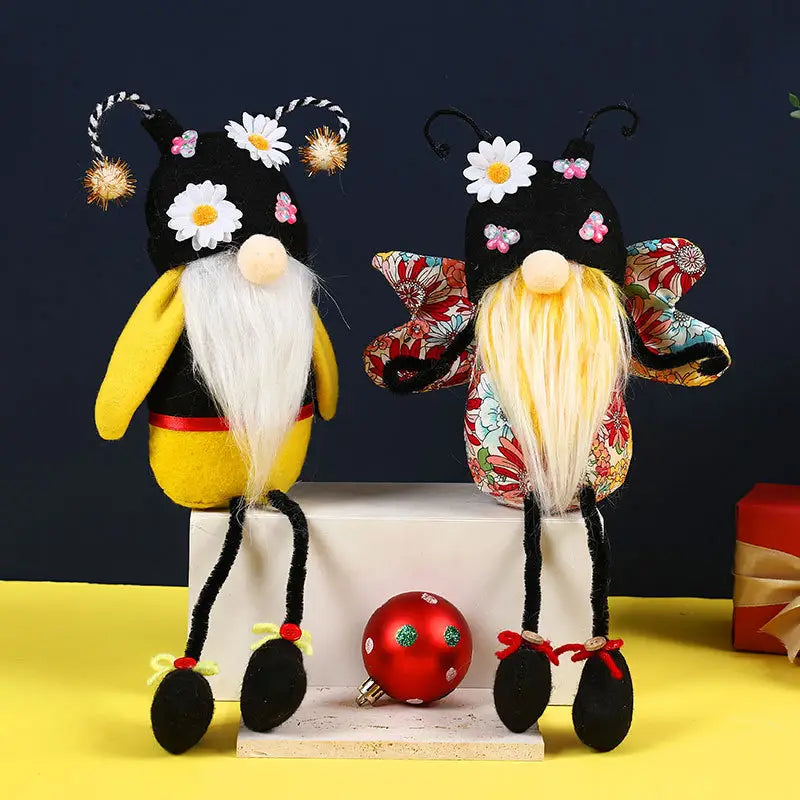 Deux décorations de peluche d’gnome abeille à longues jambes sans visage fantaisistes, comprenant l’une avec un corps jaune et blanc, une barbe blanche ornée de marguerites, et des pompons dorés sur un chapeau noir, et l’autre avec une tenue à motif floral, une barbe blonde et des fleurs assorties.