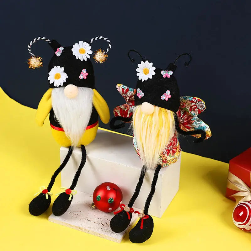 Deux peluches d’gnomes abeille à longues jambes sans visage pour décoration, avec des motifs d’abeille fantaisistes : l’une en tissu jaune et noir avec une barbe blanche, et l’autre en tissu à motif floral avec des cheveux blonds, toutes deux portant des chapeaux décoratifs ornés de marguerites et d’antenne.