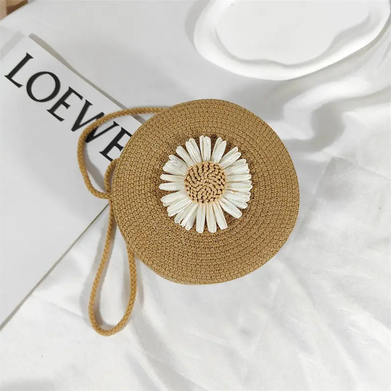 Beige woven straw circular crossbody bag adorned with a white daisy embellishment ideal as a round woven shoulder purse.