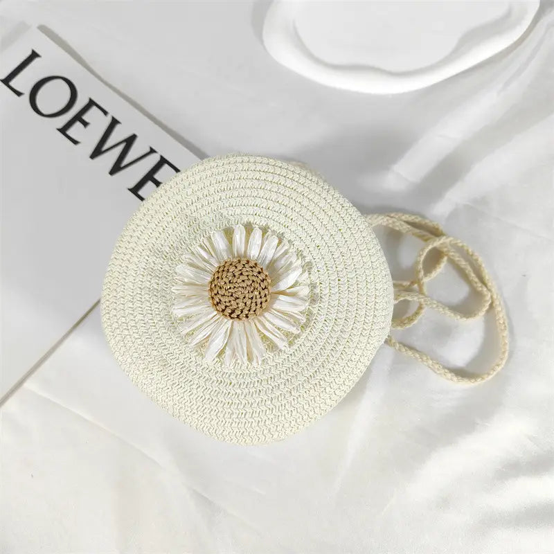 Off-white woven straw crossbody bag featuring a detailed daisy embellishment with cream petals and a tan center designed as a round woven shoulder purse.
