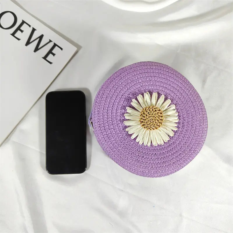 A lavender woven straw round clutch adorned with a white and beige daisy appliqué for use as a crossbody shoulder purse.