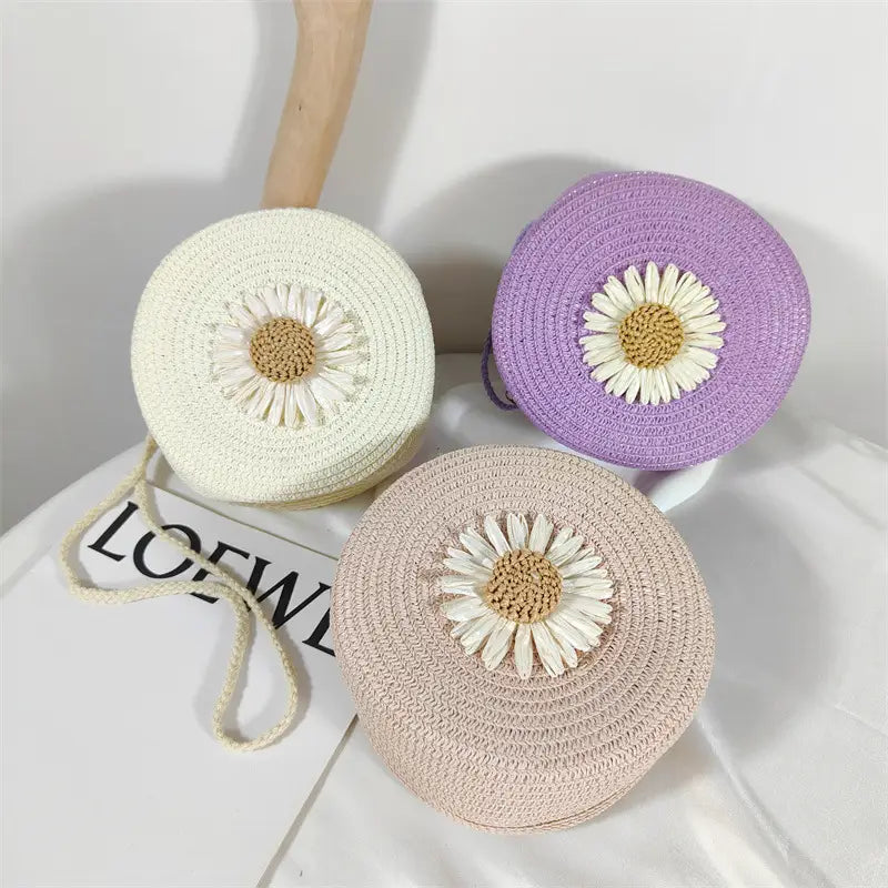 three round straw crossbody bags in cream lavender and beige each adorned with a white daisy appliqué with a tan center in the daisy straw crossbody bag round woven shoulder purse collection