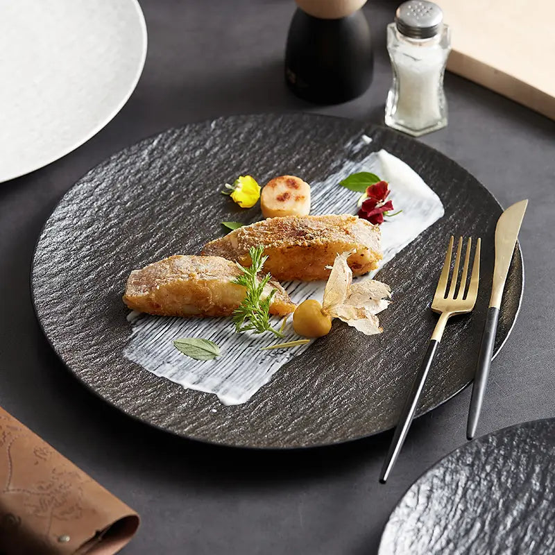 Black textured ceramic round plate with a subtle stone-like finish and a central white paper liner holding the food featuring a Nordic rock pattern.