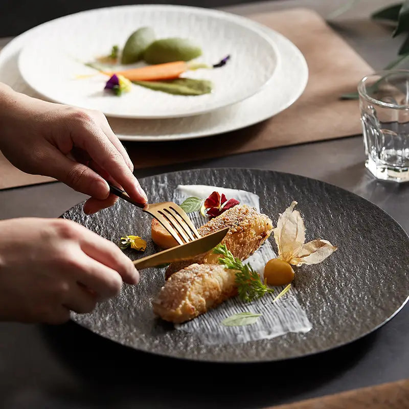 Gold-toned cutlery set resting on a dark textured ceramic round plate with Nordic rock pattern.