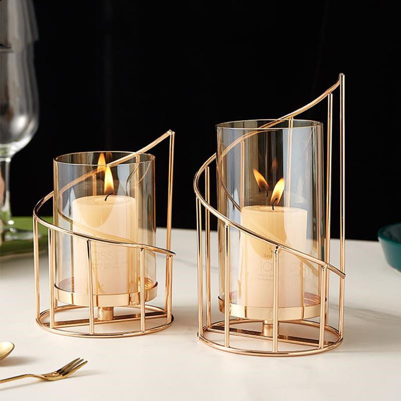 Gold metal spiral candle holder with glass cylinder features gold-toned metal holders with clear glass cylinders each cradling a lit cream-colored pillar candle.