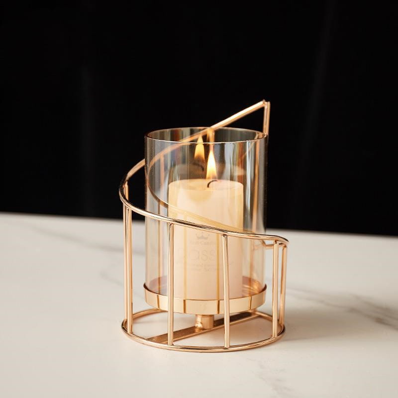 A sleek rose gold metal spiral candle holder with clear glass cylinder and curved wireframe design cradles an ivory pillar candle.
