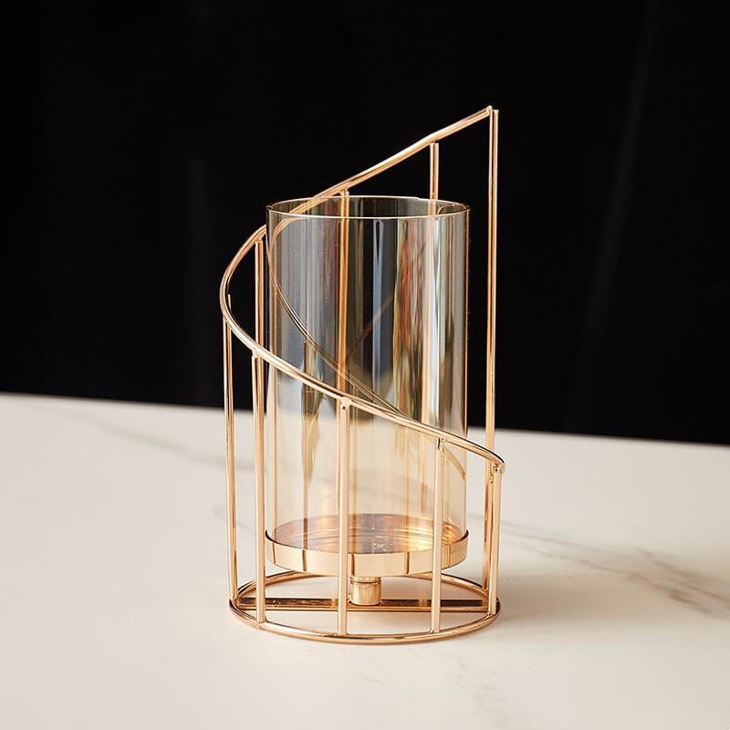 a gold metal spiral candle holder with a transparent glass cylinder encased in an elegant gold-toned metal frame featuring curved vertical bars and a circular base