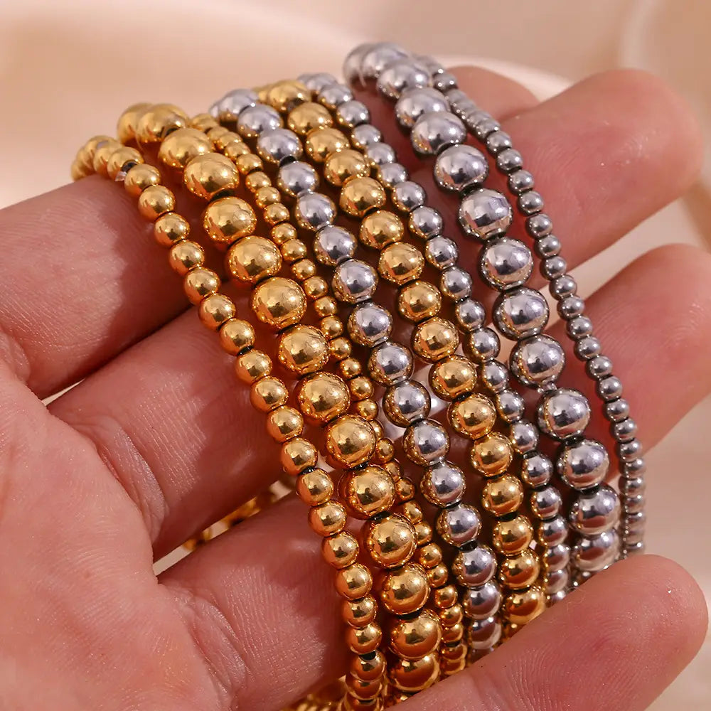 A hand holds a collection of 18k gold plated stainless steel elastic bead bracelets featuring alternating gold and silver smooth metallic spheres.