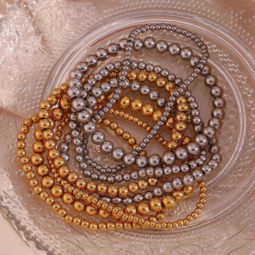 Stacks of metallic gold and silver 18k gold plated stainless steel elastic bead bracelets arranged in a circular pattern on a clear glass plate.