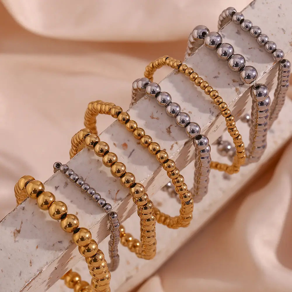 A collection of 18k gold plated stainless steel elastic bead bracelets in gold and silver tones featuring smooth spherical beads with a polished metallic finish.