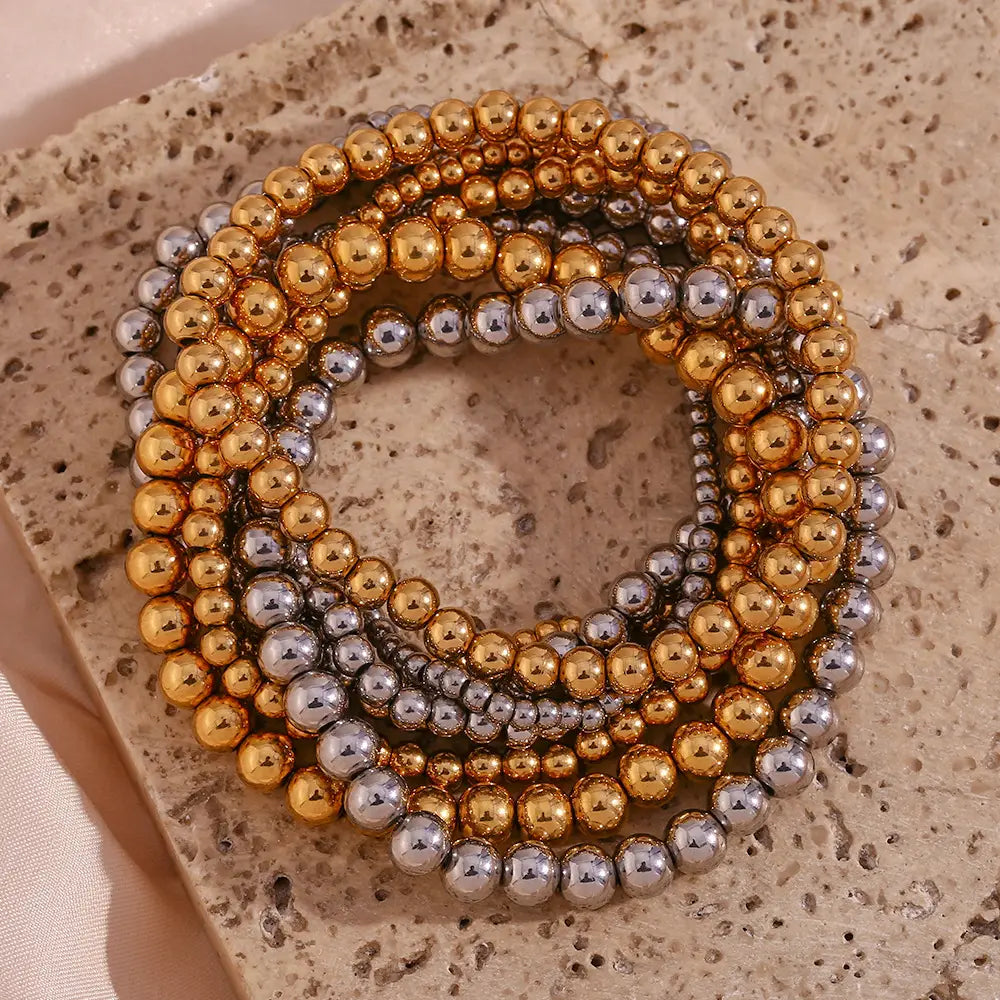 A stack of 18k gold plated stainless steel elastic bead bracelets in gold and silver tones with a glossy finish and varying bead sizes.