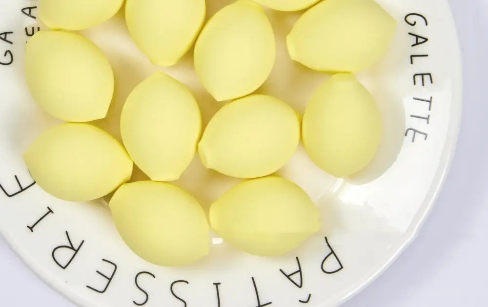 Un ensemble de mini éponges de maquillage en silicone jaune citron pâle, de forme rappelant le citron, à texture douce en forme de goutte, disposées sur une assiette blanche avec des inscriptions noires.