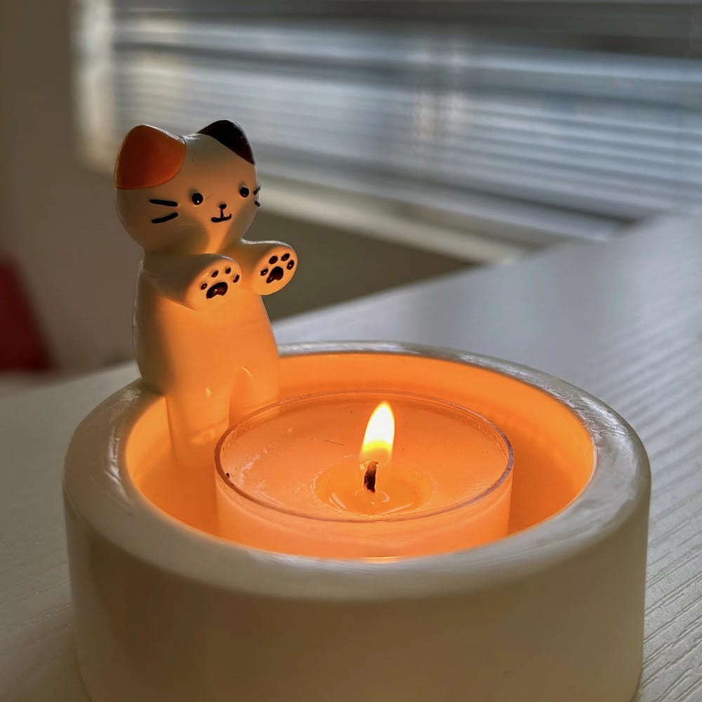 Cute cream-colored ceramic cat figurine with orange and black accents standing beside a lit tea light candle in a white holder for use as a cat candlestick holder ceramic kitten tea light stand.