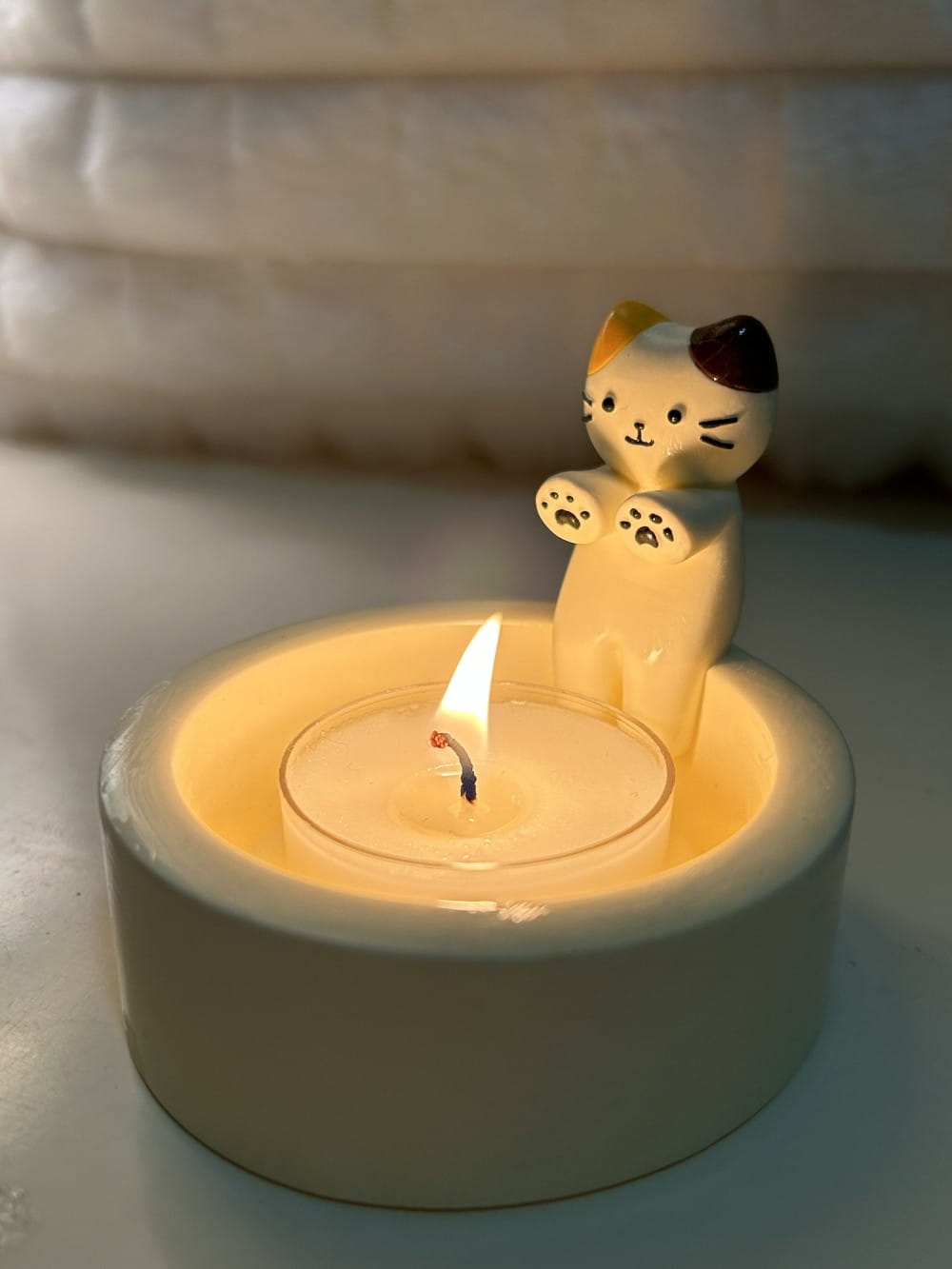 Ceramic cat candlestick holder featuring a white body with orange and black ears sitting beside a lit tea light candle in a matching ceramic holder.