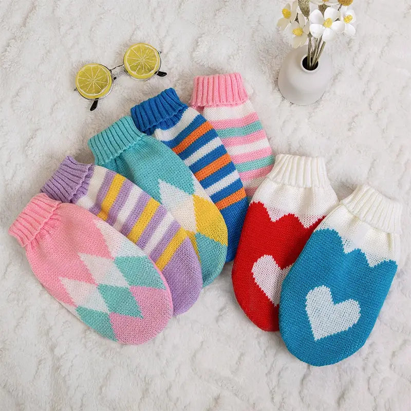 Eine Kollektion bunter gestrickter Baby-Handschuhe mit Zackenmuster- und Herzmustern, die zusammen mit einem gestrickten Hundepullover mit gestreiftem Woll-Blumenmuster für Haustiere präsentiert wird