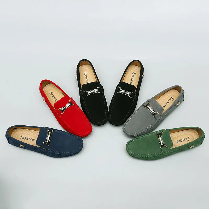 Eine Kollektion handgefertigter Herren-Slipper aus Samtleder mit Horsebit-Design als Gommino-Schuhen in Rot, Schwarz, Grau, Marineblau und Grün, jeweils mit einer silberfarbenen Metallschnalle.
