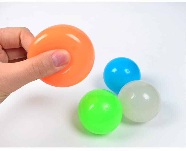 A hand holds a bright orange glossy plastic luminous sticky wall ball glow in the dark squeeze toy with three other colorful balls nearby.