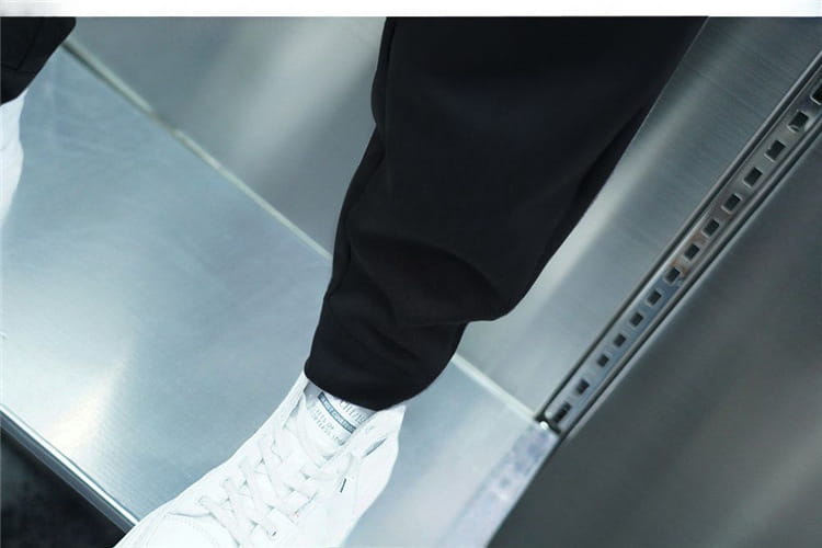 a pair of men’s casual tapered jogger pants in dark color worn inside an elevator paired with white high-top sneakers featuring black laces and a subtle logo on the tongue
