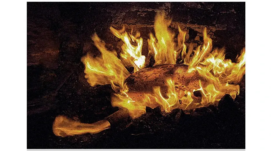 Un tronco de madera rústico de color marrón oscuro ardiendo intensamente con llamas naranja doradas en una chimenea de piedra, junto a un wok de hierro hecho a mano con asa de madera y tapa de vidrio.