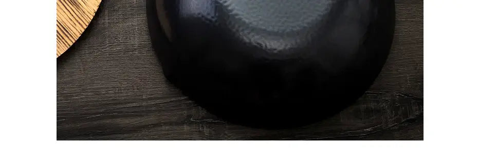 Un cuenco de cerámica negro brillante con interior liso y redondeado y discretas crestas texturizadas, acompañado de un wok de hierro hecho a mano con asa de madera y tapa de vidrio