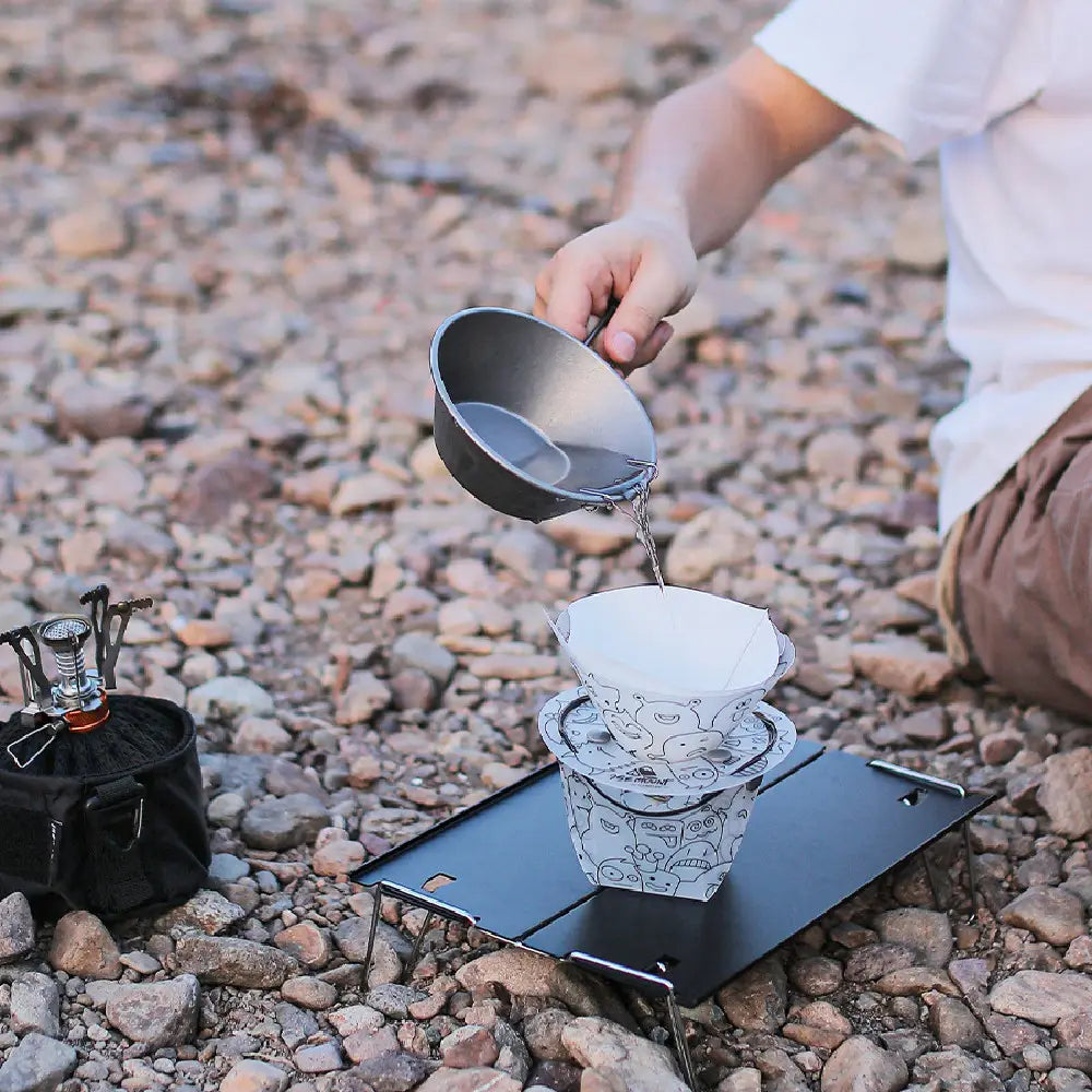 a white paper coffee filter with black cartoon illustrations resting on a stack of matching filters on a portable camping stove as part of an ultra light portable folding tableware set