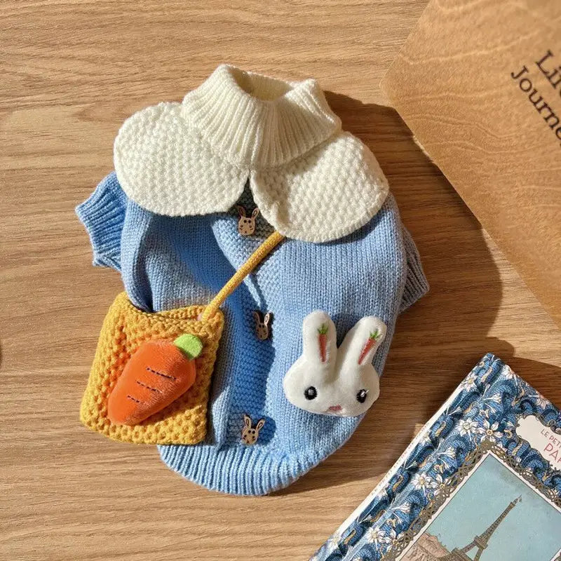Un doux pull pour animal de compagnie en tricot bleu, doté d’un col texturé contrastant de couleur crème et d’une adorable appliqué de lapin blanc avec des détails de carotte brodés.