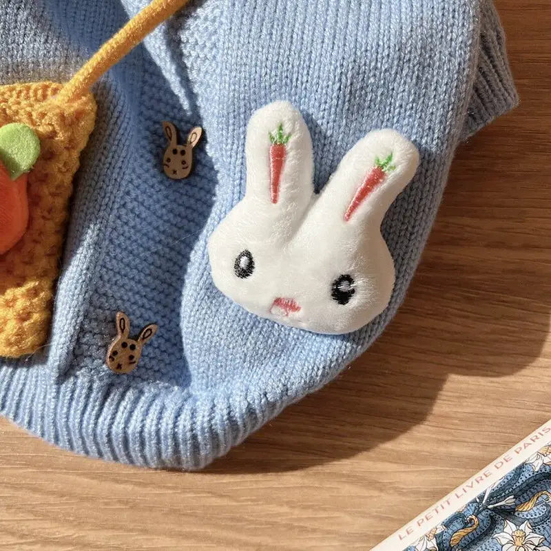 Une broche mignonne en forme de lapin blanc en peluche, ornée de carottes rouges brodées sur les oreilles et d’yeux boutonnés noirs, fixée à un pull pour animal de compagnie en tricot bleu clair.
