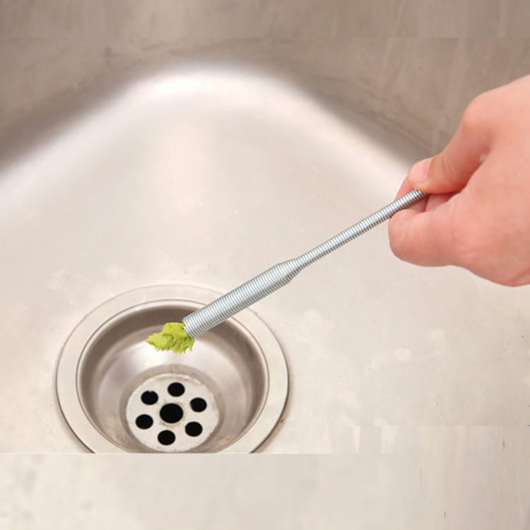 A flexible silver-colored metal drain clog remover 60cm spring pipe tool with a green fuzzy cleaning head is being used to remove debris from a sink drain.