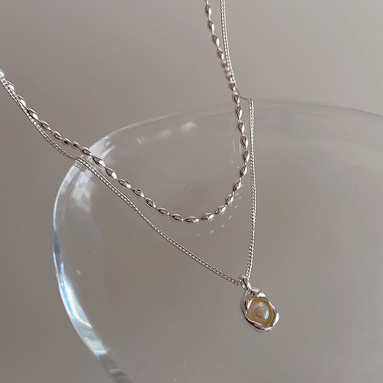 A delicate silver necklace from a set of five minimalist silver and pearl pendant necklaces featuring a twisted chain and a small teardrop-shaped pendant with an iridescent pearl-like stone.