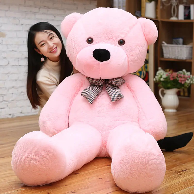 A plush pink teddy bear stuffed animal with a black nose dark eyes and a checkered bow tie designed as a toy for kids available in sizes from 40 to 200 centimeters.