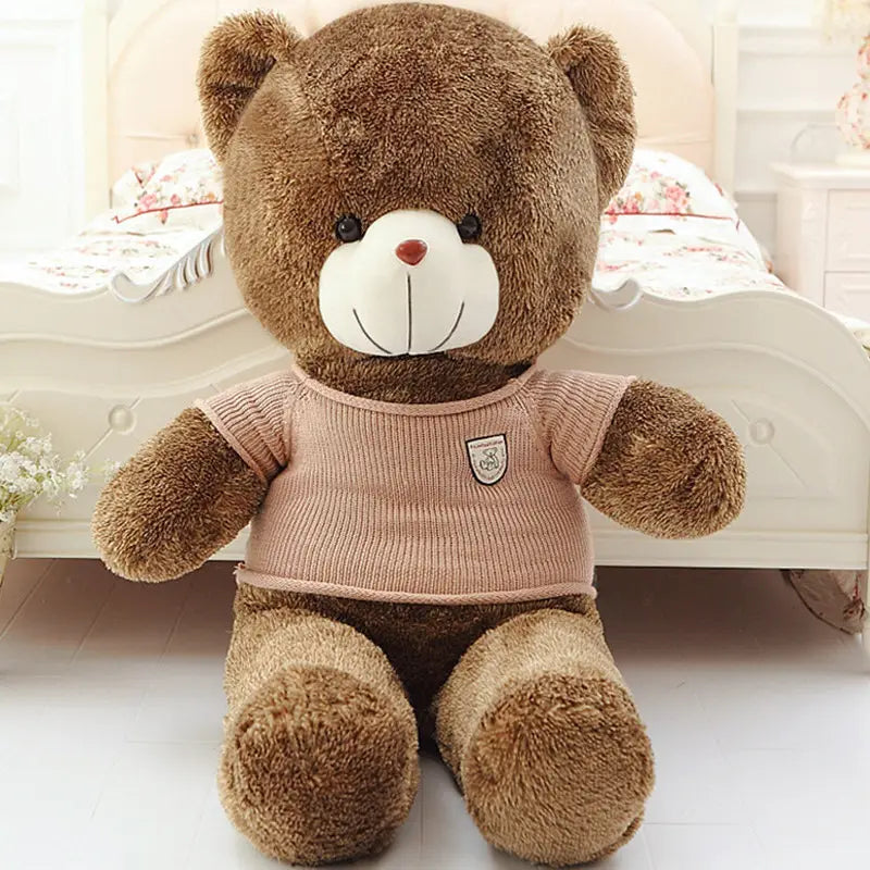 A brown plush teddy bear wearing a pink ribbed knit shirt with a small shield-shaped patch on the chest perfect as a stuffed animal toy for kids sized 40-200cm.