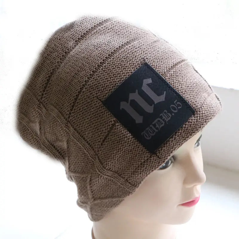 Beige knitted men’s fleece lined winter beanie hat featuring a black rectangular patch with gray lettering.