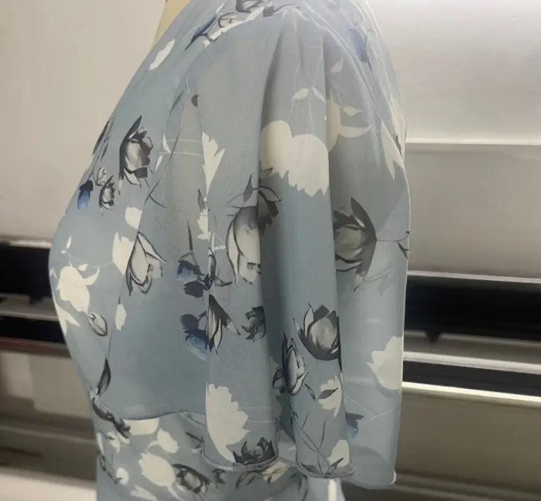 Une robe longue d’été à décolleté en V floral volanté bleu clair, avec un tissu doux et fluide et de délicats motifs de fleurs noires et blanches.