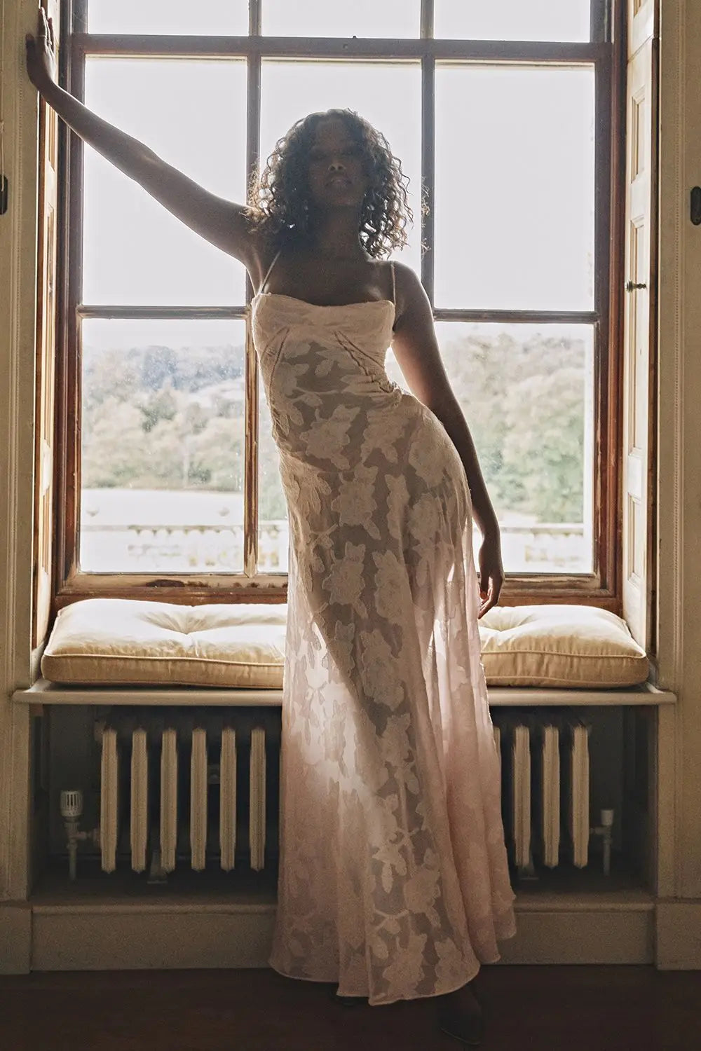 Une robe de soirée longue maxi sans dos en dentelle florale blanc cassé, fluide, avec de délicats motifs floraux et de fines bretelles spaghetti.