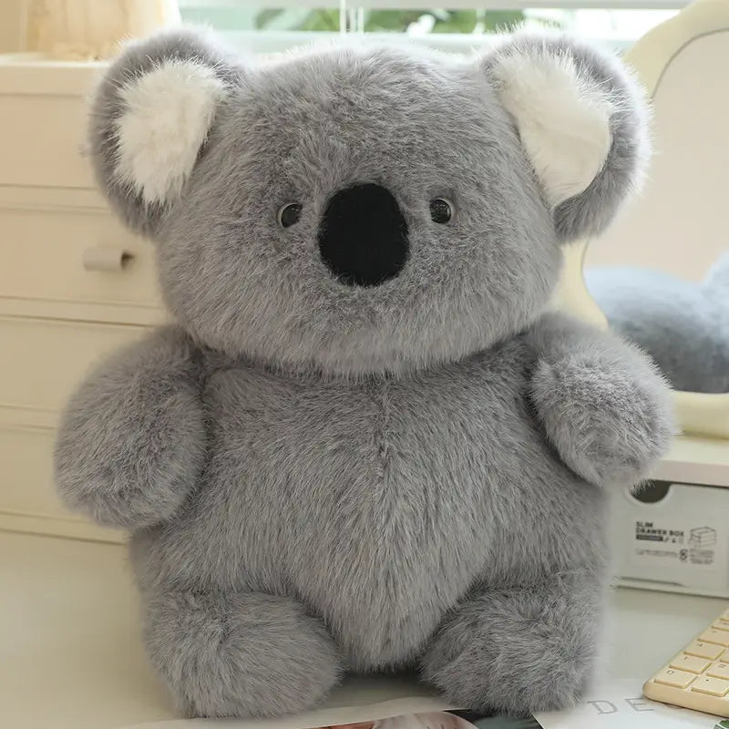 平らに寝かせた、もっちりしたグレーのコアラのぬいぐるみで、やわらかなふわふわの毛並みと、ぬいぐるみ素材でできた白い内側の耳を備えたもの。