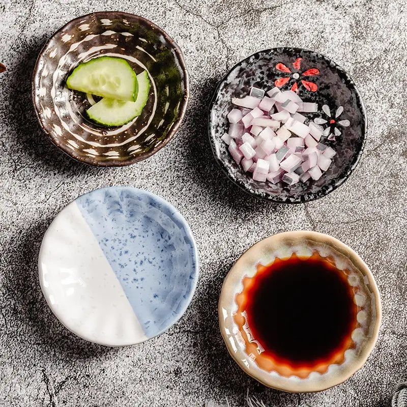 Quatre bols à condiments en céramique, peints à la main avec des motifs uniques : bordure cannelée brune et blanche, motif floral rouge et noir, motif bleu et blanc moucheté, et bord beige cannelé ; chacun contient une sauce soja foncée pour servir des en-cas.